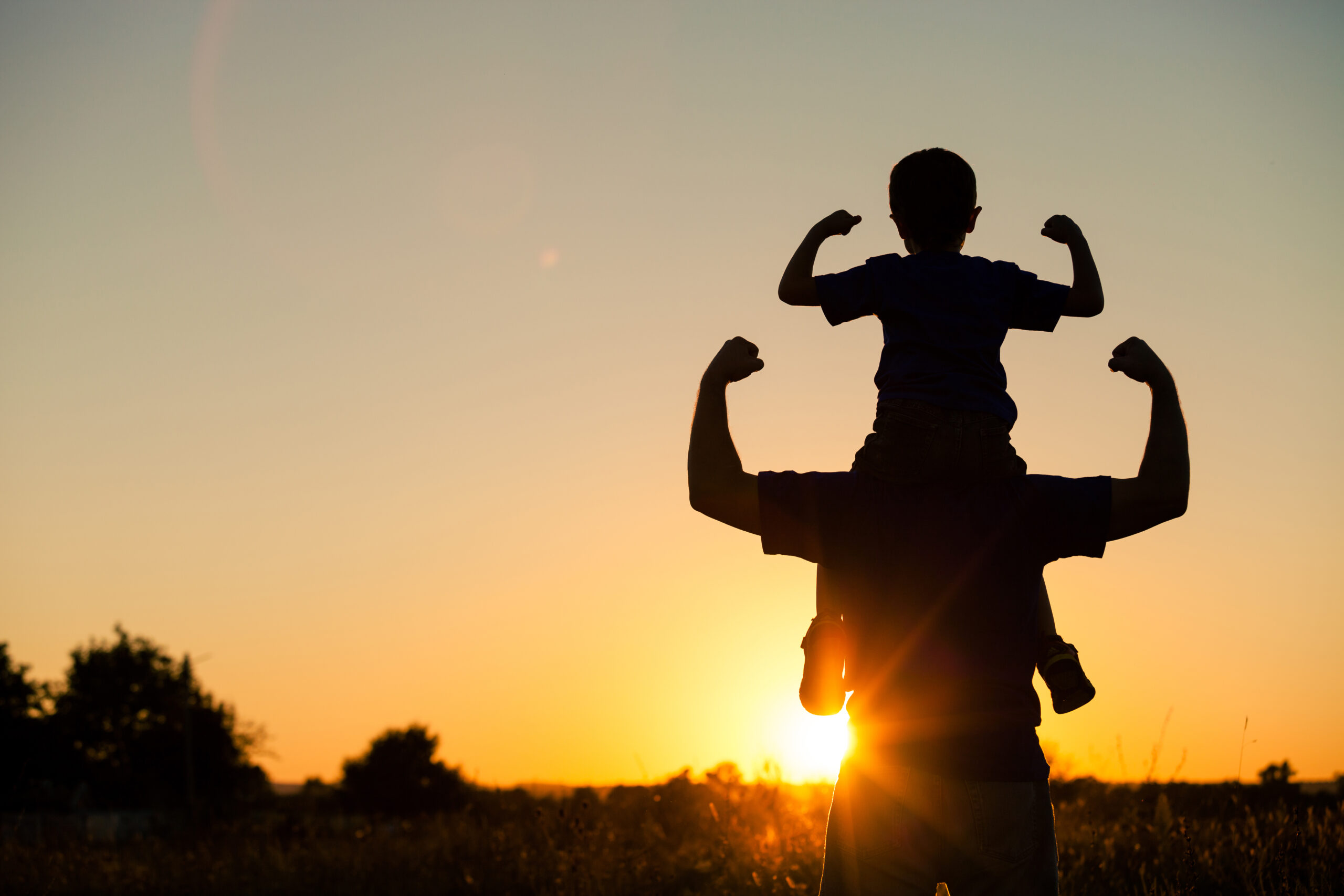 Father And Son Playing In The Park At The Sunset Time. man holding son on shoulders, both flexing, silhouette, sun in background.