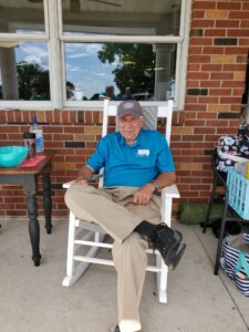 Carl sitting in a rocking chair with a hat on that says "loving life."